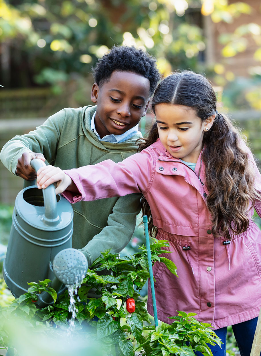 Two children — boy and girl — hold a watering can together and water a garden